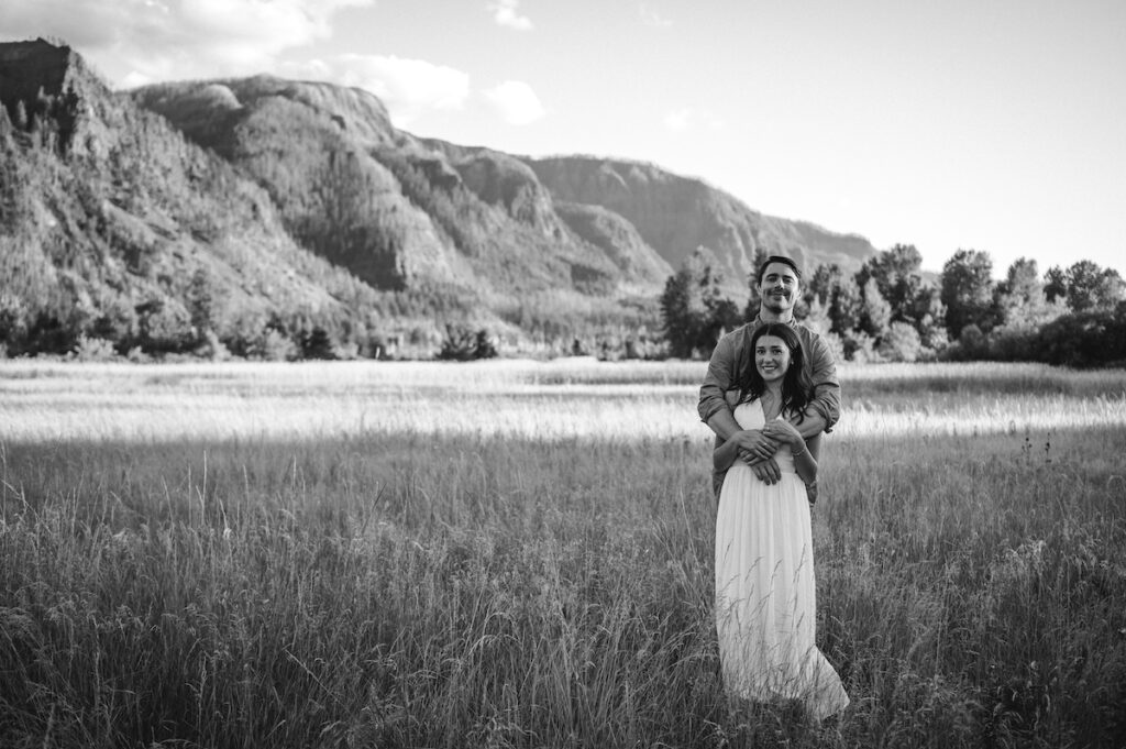 black and white image of couple standing in field with columbia river gorge behind them 