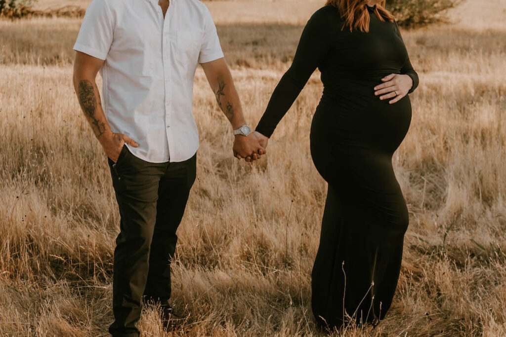 Husband and wife hold hands in golden field during sunset maternity session in Oregon